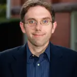 A man with short brown hair and glasses in a dark suit and striped shirt stands in front of a brick building, smiling at the camera.