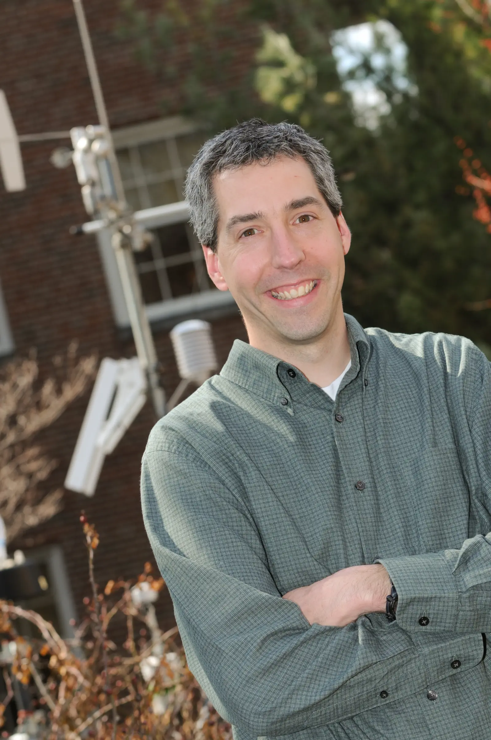 A man with short gray hair and a green button-up shirt stands outdoors with arms crossed, smiling. He is in front of a brick building and some equipment with greenery in the background.