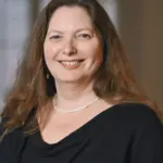 A smiling woman with long brown hair wearing a black top and a pearl necklace stands in front of a blurred background.
