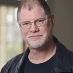 A man with short hair and glasses, wearing a black leather jacket over a black shirt, smiles softly. He has a trimmed beard and stands in front of a window with a soft, blurred background.