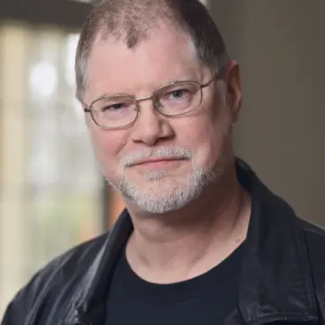 A man with short hair and glasses, wearing a black leather jacket over a black shirt, smiles softly. He has a trimmed beard and stands in front of a window with a soft, blurred background.