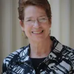 A smiling woman with short, brown hair wearing glasses and a patterned black and white jacket stands in front of a neutral background.