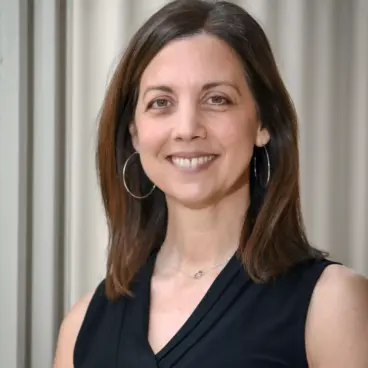 A woman with shoulder-length brown hair is smiling while standing in front of a neutral background. She is wearing a black sleeveless top and hoop earrings.