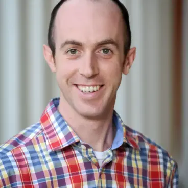 A person with short hair is smiling at the camera, wearing a plaid shirt with red, yellow, and blue stripes. The background is blurred, featuring vertical lines.