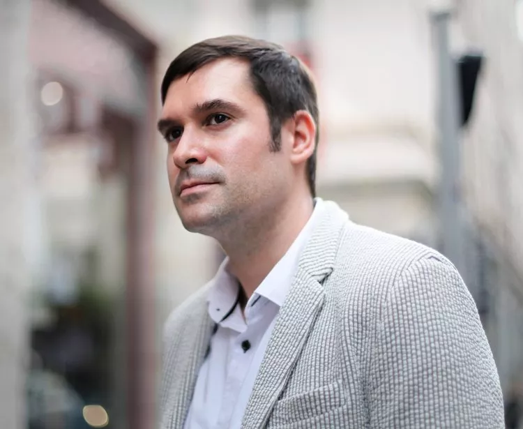 A man in a light gray blazer and white shirt stands outdoors, gazing thoughtfully into the distance. The background is softly blurred, suggesting an urban environment.
