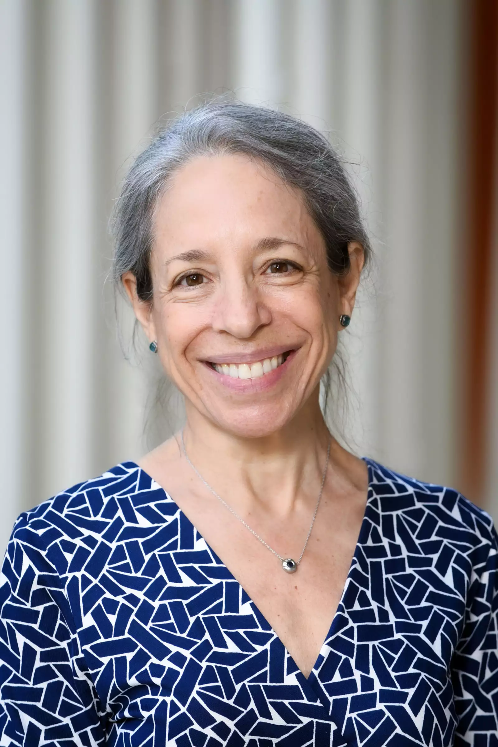A woman with gray hair, smiling, wearing a blue patterned dress and a silver necklace. She is standing in front of a blurred light-colored background.