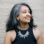 A woman with long dark and gray hair smiles while looking to the side. She is wearing a black sleeveless top, geometric earrings, and a statement necklace with vertical metal bars. The background is a light-colored brick wall.