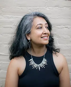 A woman with long dark and gray hair smiles while looking to the side. She is wearing a black sleeveless top, geometric earrings, and a statement necklace with vertical metal bars. The background is a light-colored brick wall.