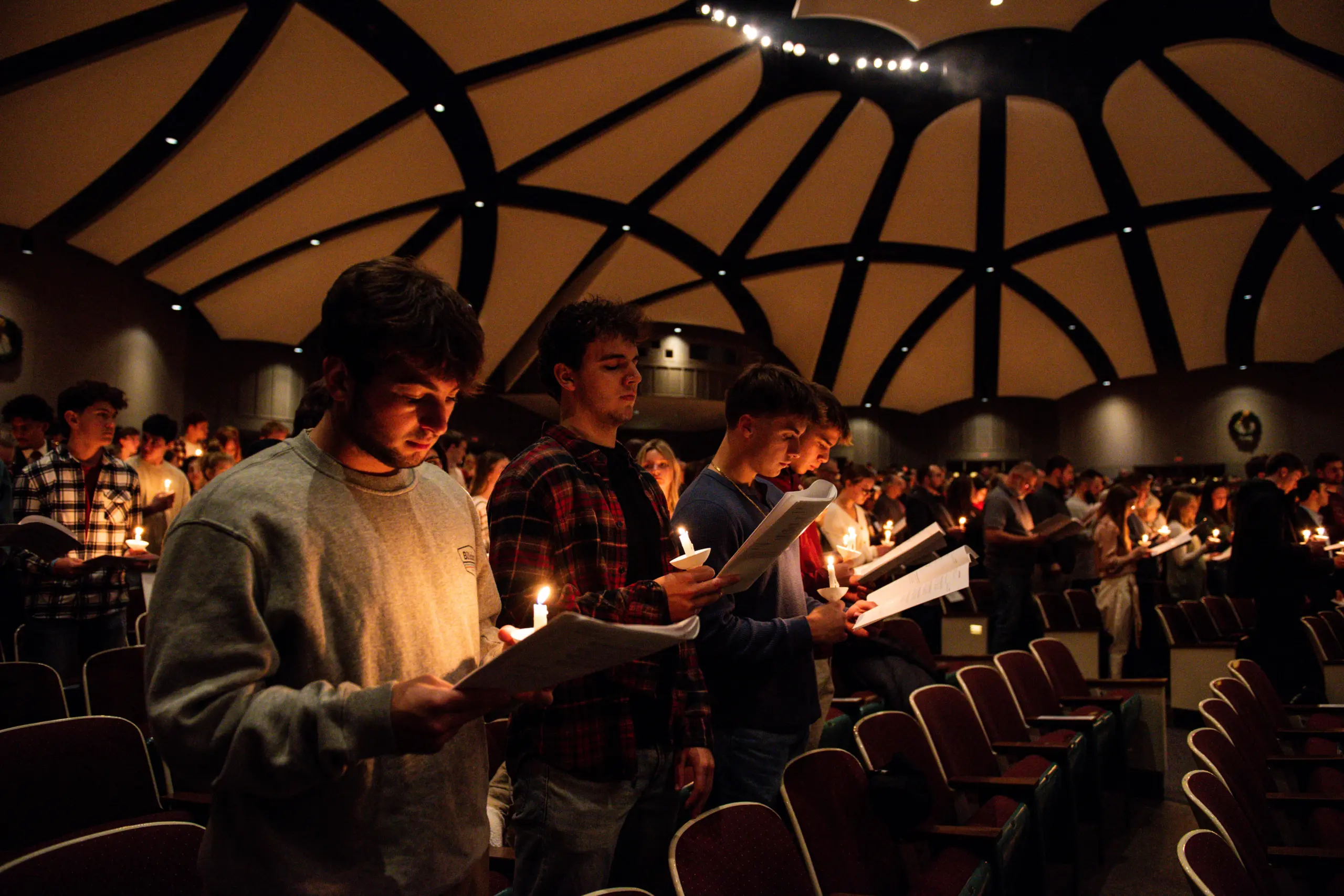 People standing in a dimly lit auditorium, each holding a lit candle and a program or song sheet. The ceiling has a unique pattern of curved beams. The atmosphere is solemn and reflective, suggesting a ceremony or memorial.