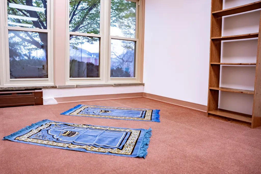 A prayer room with two blue prayer rugs on a red carpeted floor. There are large windows with trees visible outside and an empty wooden shelf on the right side.