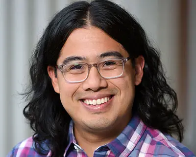 A person with long, dark hair and glasses smiles at the camera. They are wearing a plaid shirt and the background is softly blurred.