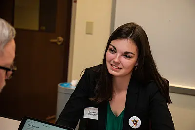 A woman with long dark hair wearing a black blazer and name tag is sitting at a table in a meeting room. She is speaking to a man across from her who is partially visible. There is a whiteboard and door in the background.