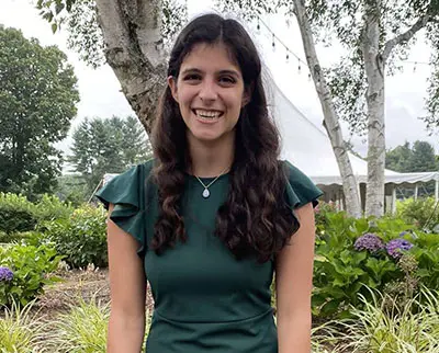 A woman with long dark hair smiles while standing in a garden. She is wearing a green dress, and there are trees and purple flowers in the background.