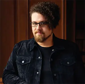 A person with curly hair and a beard is standing indoors. They are wearing glasses and a dark jacket over a black shirt. The background features wooden paneling.