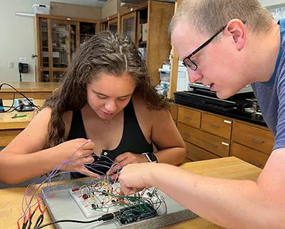 Two people work on a circuit board with various wires in a classroom setting. One is a young woman with long hair, focused on connecting wires. The other is a man with glasses assisting her. Classroom equipment is visible in the background.