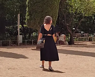 A person in a black dress walks through a park on a sunny day, carrying a bag. Trees and benches are visible in the background.