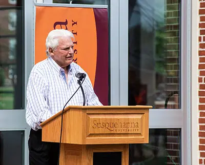 An elderly man with white hair speaks at a wooden podium with "Susquehanna University" inscribed on it. He stands in front of a banner and glass window, wearing a striped shirt.
