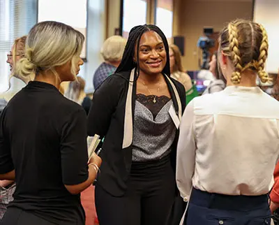 A group of professionally dressed people engage in conversation at a networking event. The setting appears to be a conference room with large windows.