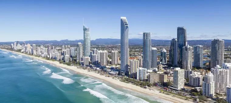 Aerial view of a coastal city skyline with tall skyscrapers lining the beach. The ocean waves roll toward a sandy shore, and the cityscape extends into the distance under a clear blue sky.