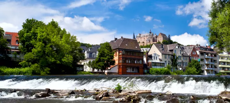 A scenic view of a river with a small waterfall in the foreground. Behind it, a row of colorful houses sits under a clear blue sky. On a hill in the background, a historic castle overlooks the scene surrounded by lush greenery.