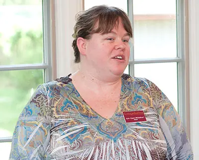 A woman with short brown hair is speaking indoors. She wears a colorful, patterned blouse and has a name tag. The background features large windows showing greenery outside.