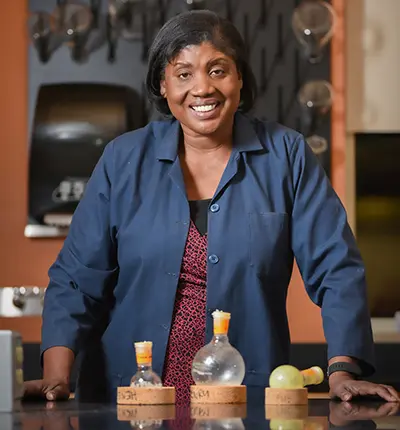 A person smiling and standing behind a table with laboratory glassware. They wear a blue lab coat over a red patterned shirt. The background shows a wall with hanging lab equipment.