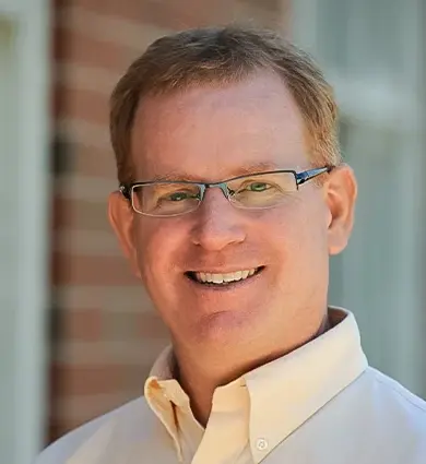 Smiling person wearing glasses and a light-colored button-up shirt, standing in front of a brick wall with windows.