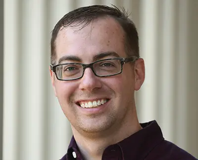 Smiling person wearing glasses and a dark shirt stands in front of a light-colored background with vertical lines.