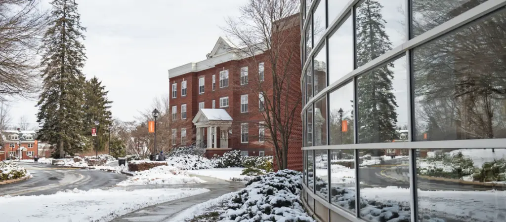 Snow-covered college campus with a historic red-brick building and a modern glass building. Snow-dusted trees and shrubs line the curved road, and orange banners hang on lamp posts. The glass building reflects the wintery scene.