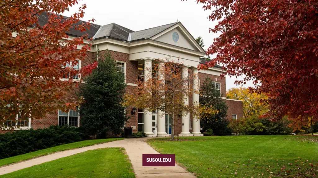 A large brick building with white columns is surrounded by green grass and trees with red and yellow autumn leaves. A pathway leads to the entrance. A sign reading "SUSQU.EDU" is in front of the building.