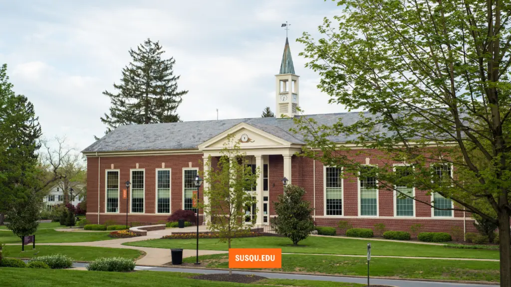 A red brick building with a clock tower and white columns stands surrounded by green lawns and trees. It features large windows and a neatly landscaped garden. A sign in front reads "SUSQU.EDU.