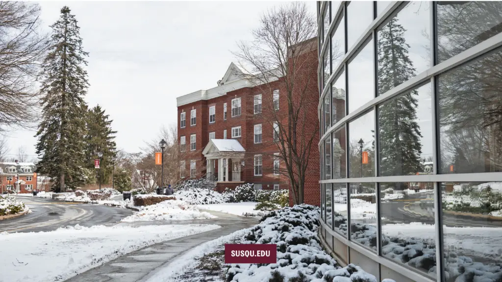 Snowy campus scene with a red brick building in the background featuring a white-columned entrance. A modern glass building with reflections is on the right. Snow covers the ground and trees, and there is a sign labeled "SUSQU.EDU.
