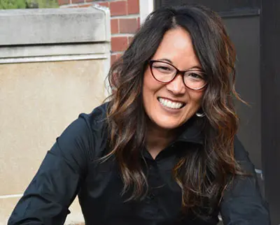 A smiling person with long, wavy dark hair and glasses is wearing a black shirt and sitting outdoors against a brick and stone wall.