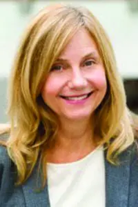 A woman with shoulder-length blonde hair smiles at the camera. She is wearing a gray blazer over a light-colored top, and the background is softly blurred.