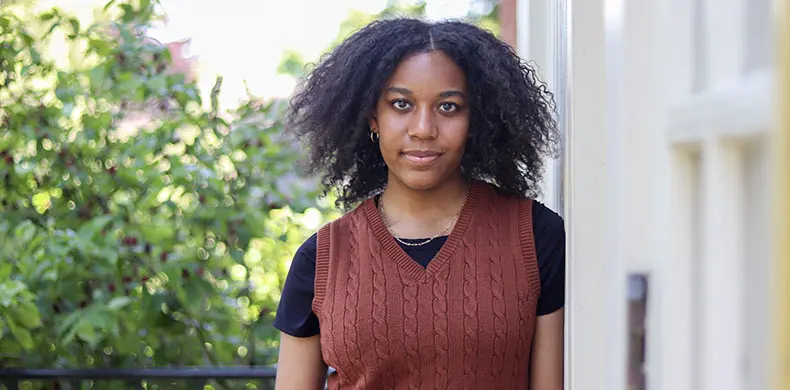 A person with curly hair stands outdoors, leaning against a light-colored wall. They are wearing a brown cable-knit vest over a black shirt. Green foliage is visible in the background.