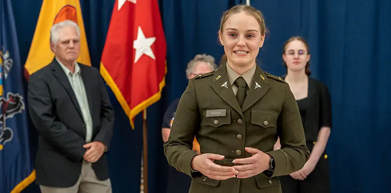 A young woman in a military uniform stands in front of three people, delivering a speech. Two flags are visible in the background, including one with red and white stars. The setting appears formal.