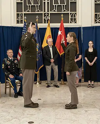 A military officer swears in a new recruit in a formal ceremony. They stand facing each other with hands raised. Flags and a blue curtain are in the background. Observers, including military personnel, watch the ceremony.