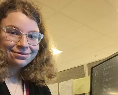A person with curly hair, wearing glasses, smiles while seated at a desk. They have a computer screen displaying code in front of them, and there are papers on a partition wall in the background.