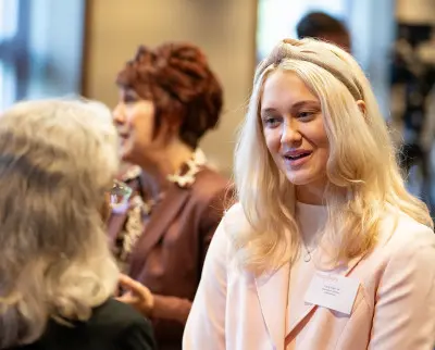 A woman with long blonde hair and a headband is engaged in conversation with another person. She is smiling and wearing a name tag. In the background, two other people are talking, with one gesturing expressively. The setting appears to be a conference or event.
