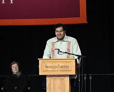 A person stands at a podium with "Susquehanna University" written on it, speaking into a microphone. They are wearing a light-colored shirt with an embroidered pattern. In the background, there is another person seated.