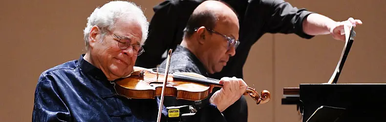 A violinist plays passionately with eyes closed, wearing a dark shirt. Behind him, another musician in glasses and a dark shirt plays the piano, focused on the keys. A third person stands in the background, partially visible.