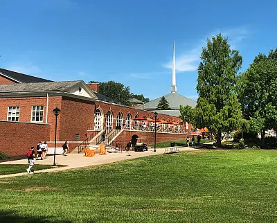 A brick building with arched windows and stairs leads to a grassy area under a clear blue sky. A tall spire rises from a nearby structure. People are walking on the path, and orange chairs are scattered along the walkway.