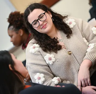 A woman with curly hair and glasses, wearing a beige cardigan with flower patterns, is engaged in a conversation with another person. They are seated in a classroom setting, with others visible in the background.