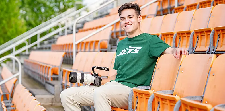 A person in a green Jets shirt sits in empty stadium seats, smiling and holding a camera with a large zoom lens. The seating is orange, and trees are visible in the background.