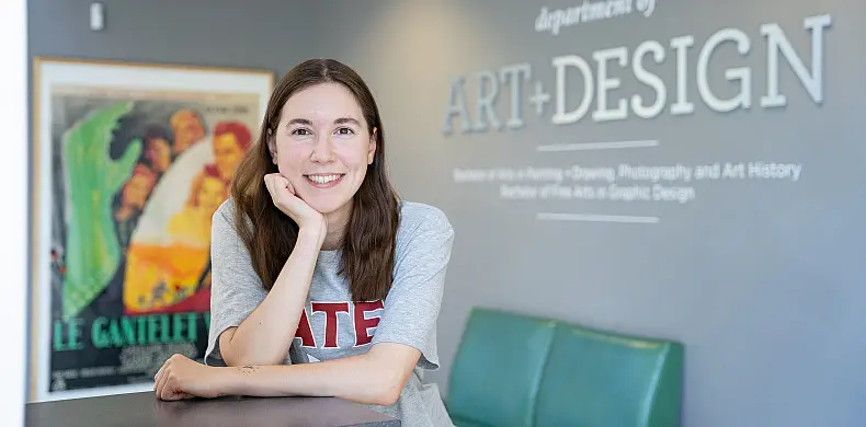 A person smiling, leaning on a counter in front of a wall with "Department of Art + Design" signage. They are wearing a gray shirt with "State" printed on it. A colorful poster is partially visible on the wall.