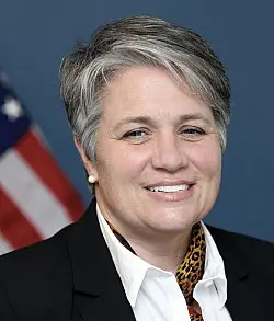 A person with short gray hair, wearing a black blazer and white shirt with a leopard print detail, is smiling in front of a blurred American flag backdrop.