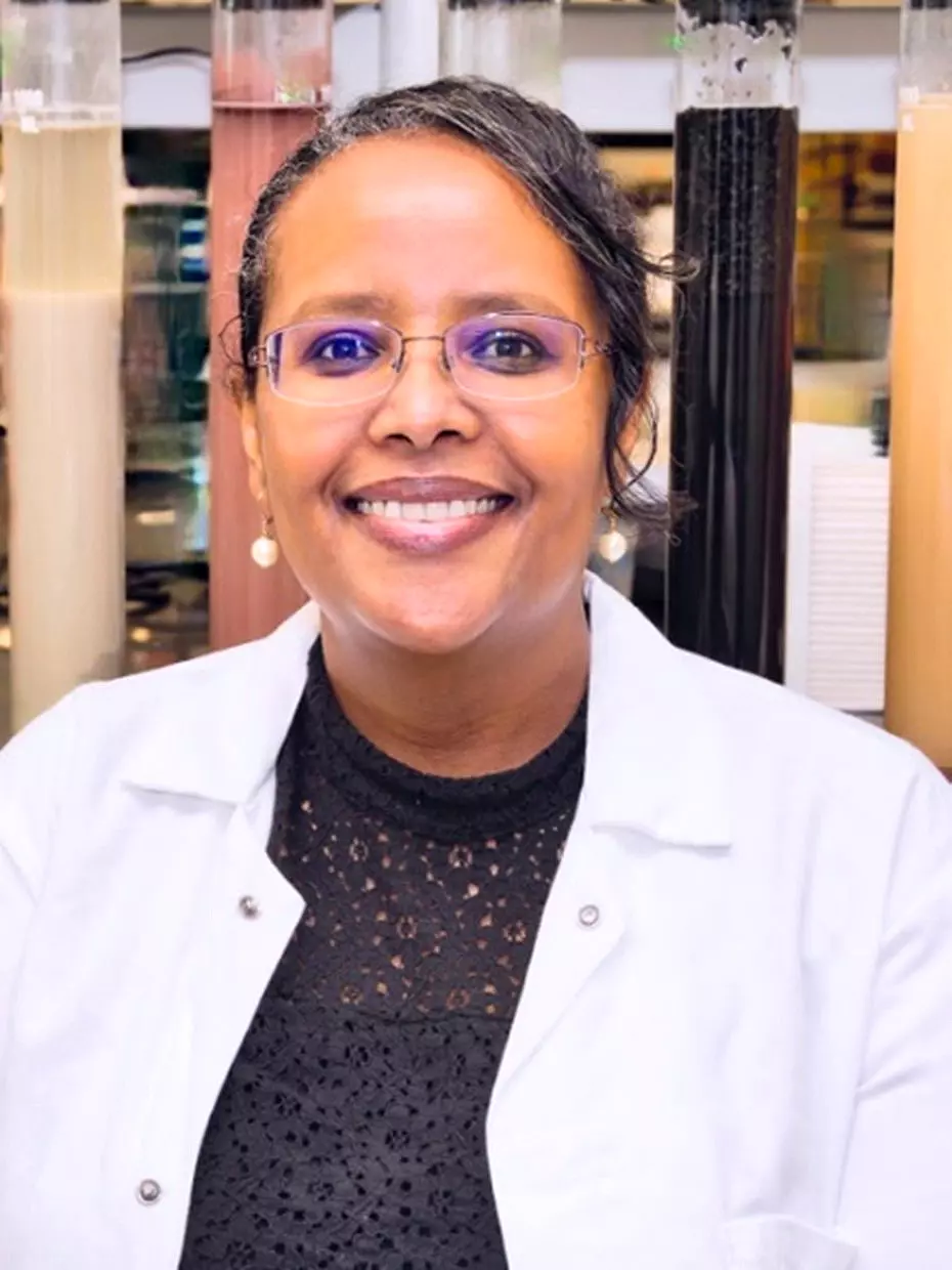 A person smiling, wearing glasses and a white lab coat. They stand in front of tall, multicolored containers, suggesting a scientific or laboratory setting.