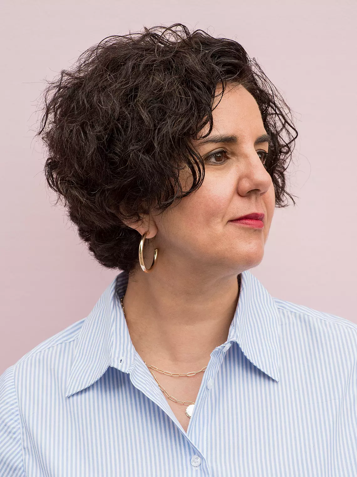 A woman with short, curly dark hair wearing hoop earrings and a blue striped shirt looks to the side against a soft pink background.
