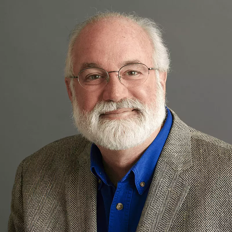 A man with glasses and a white beard smiles slightly. He wears a gray patterned blazer over a blue shirt. The background is plain gray.