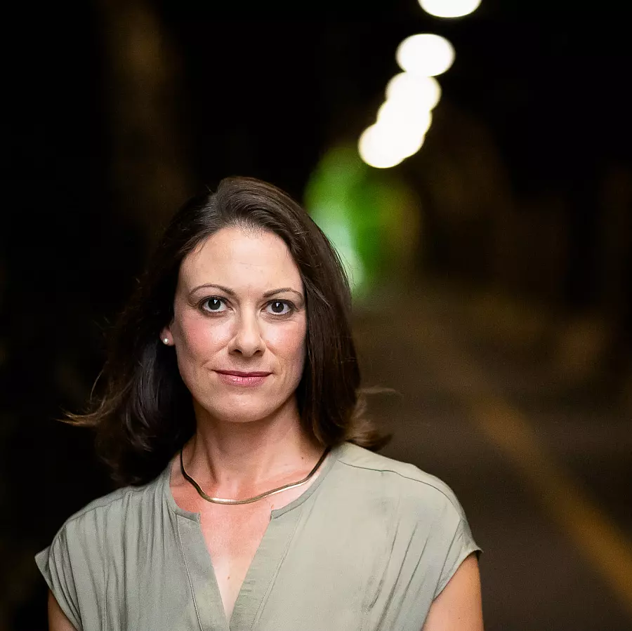 A woman with shoulder-length brown hair stands in focus against a blurred, well-lit tunnel. She wears a light gray blouse and a subtle necklace, looking directly at the camera with a neutral expression.